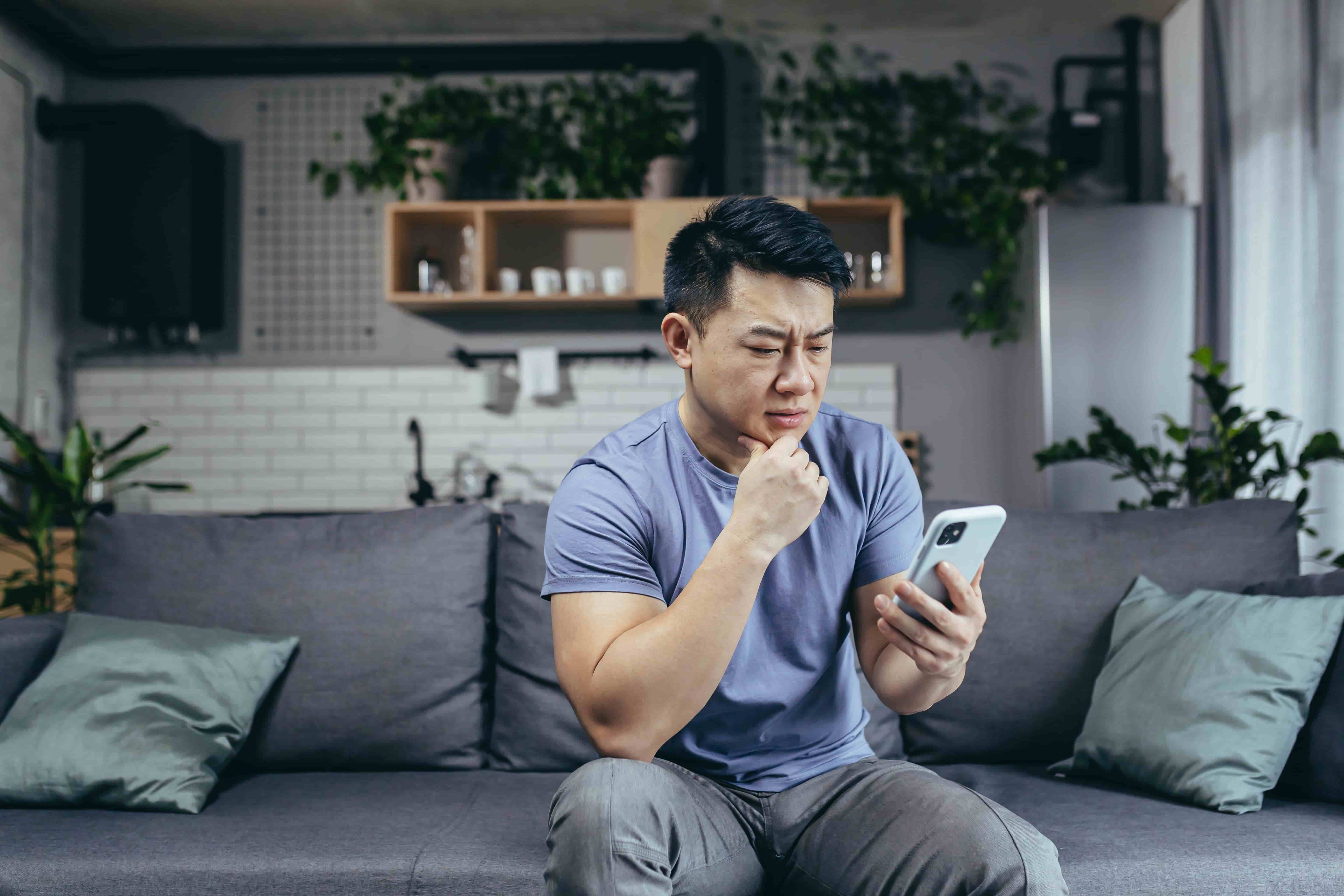 Concerned man reading news online looking at phone screen, Asian sitting on sofa at home, serious and sad